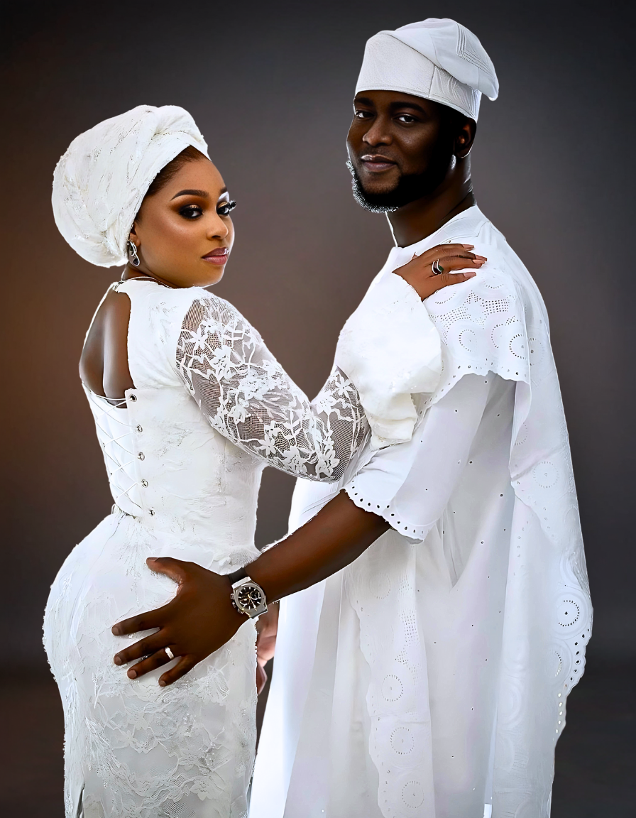 A luxurious couple dressed in traditional wedding attire. The bride wears a stunning white skirt and blouse with intricate detailing, while the groom is dressed in a matching white agbada.