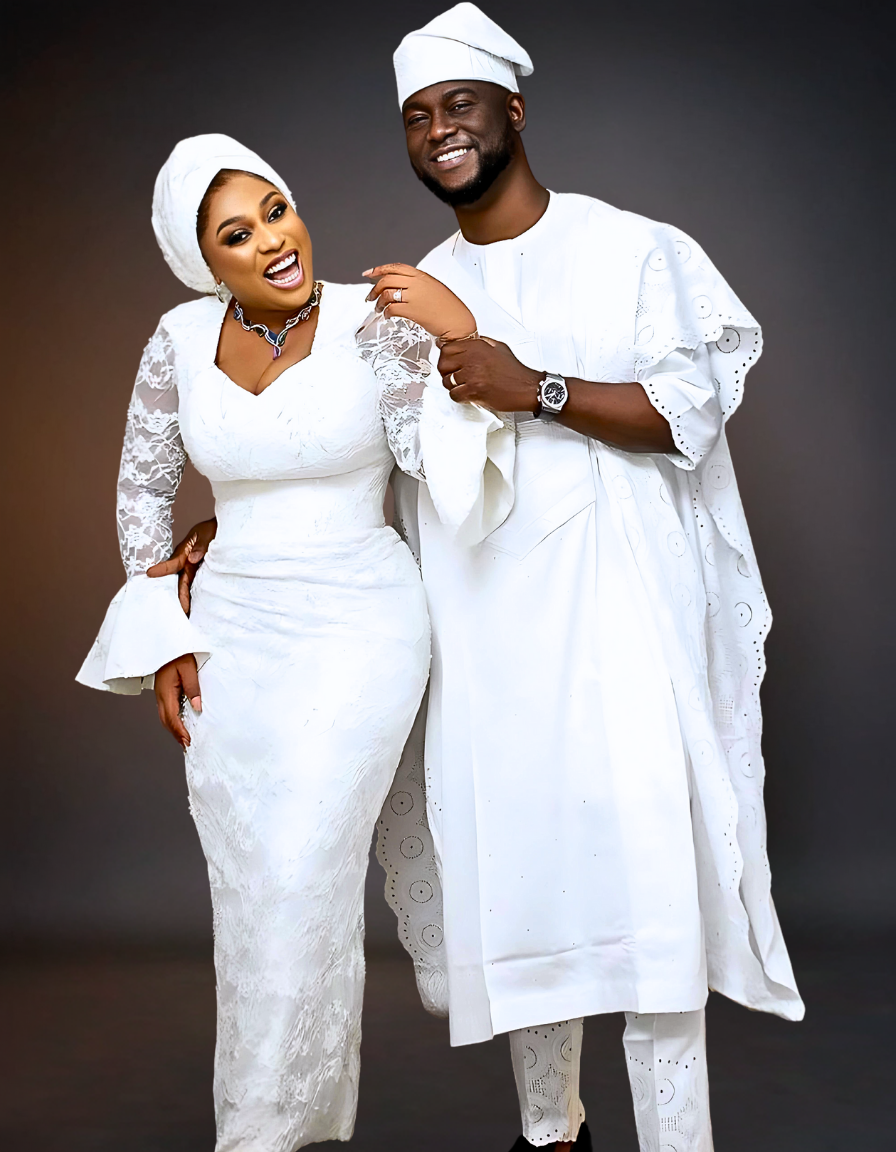 A luxurious couple dressed in traditional wedding attire. The bride wears a stunning white skirt and blouse with intricate detailing, while the groom is dressed in a matching white agbada.