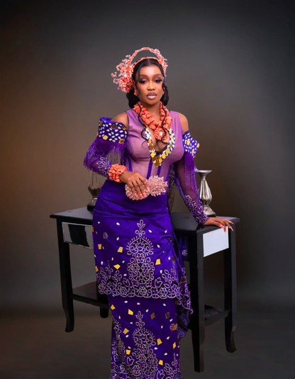 Woman in a traditional outfit with intricate designs and jewelry, standing against a dark background.